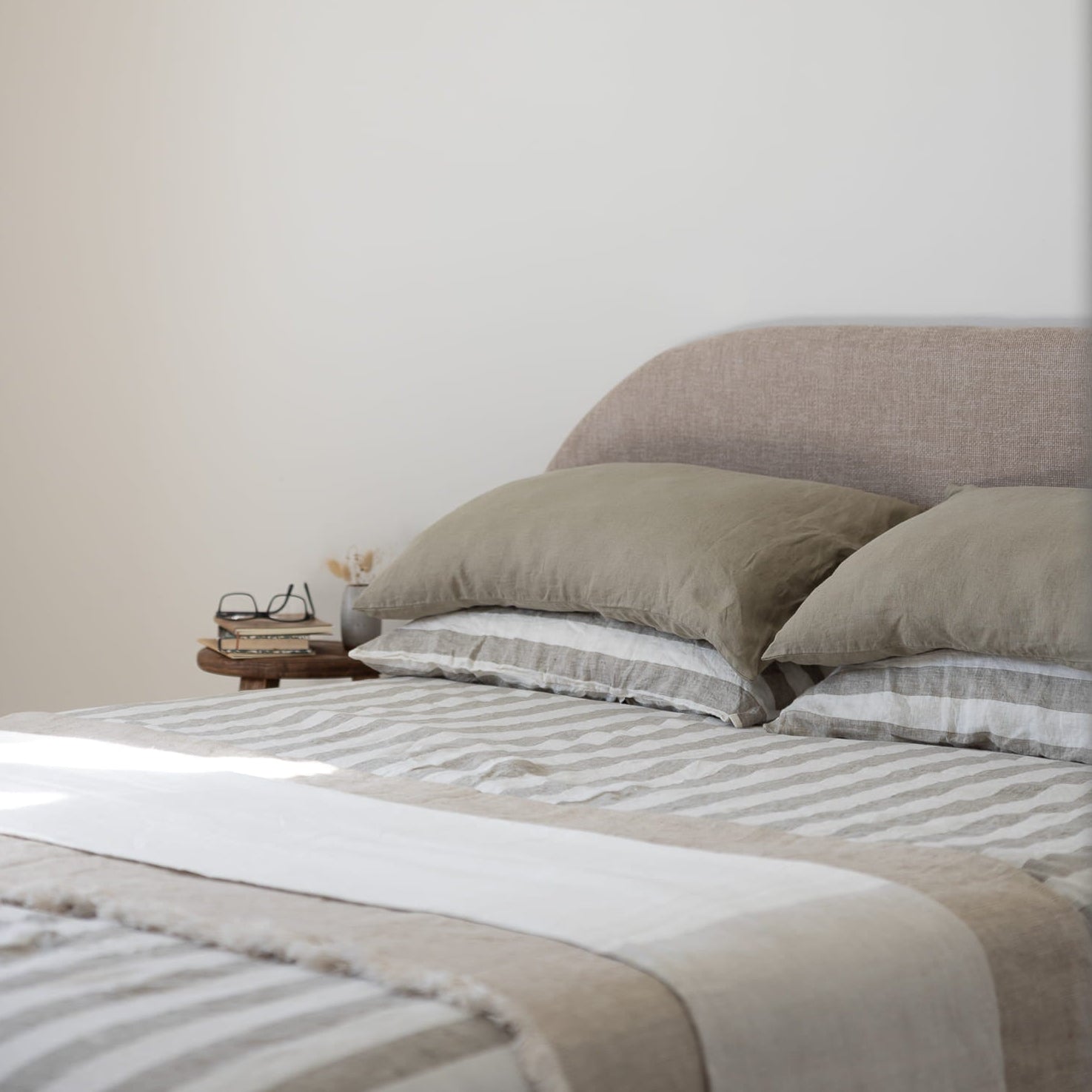 A minimalist bedroom scene featuring a neatly made bed with moss green and white striped linen bedding, matching pillows, and a textured beige headboard against a warm off-white wall.