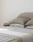 A minimalist bedroom scene featuring a neatly made bed with moss green and white striped linen bedding, matching pillows, and a textured beige headboard against a warm off-white wall.